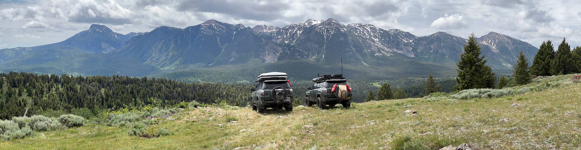 Panorama of two overland 4x4s parked on a high grass ridge, a long snow-streaked mountain range running the full width of the horizon behind them