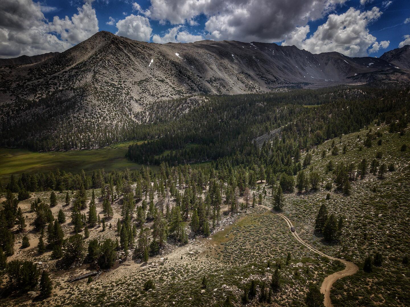 Aerial view of a high alpine basin, a dirt track winding through pine and sage toward a granite ridgeline