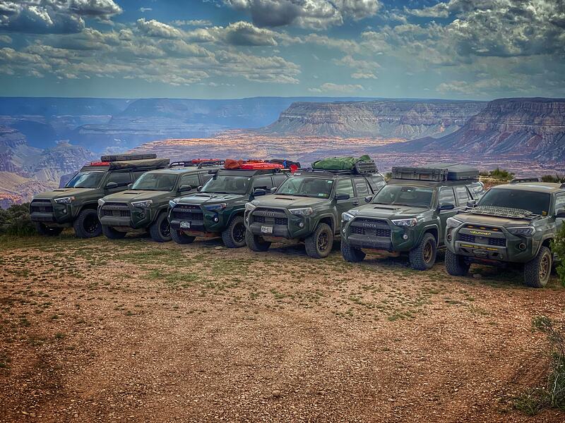 Six overland 4x4s lined up on a gravel rim, a vast canyon system stretching to the horizon behind them