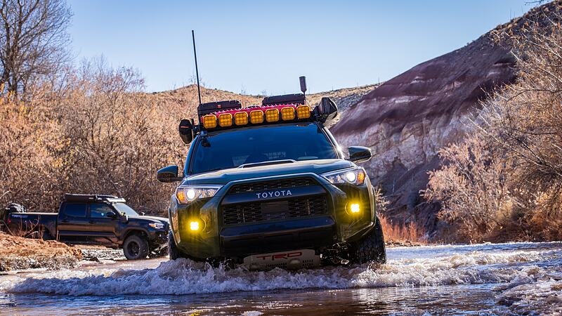 A 4x4 fording a creek in winter high desert, water breaking over the front bumper, a second truck waiting on the bank behind