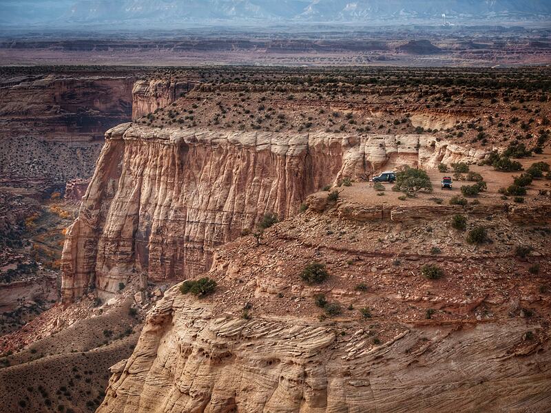 Aerial view of two 4x4s camped at the edge of a sandstone cliff, a desert canyon dropping away beneath them